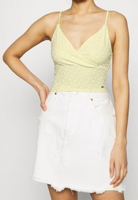 Woman wearing a light green floral spaghetti strap top and a white denim skirt with frayed hem, hand resting on hip.