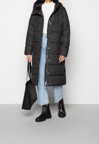 Sort pufferjakke med hætte, med en quiltet design, lynlås og sidelommer. Kombineret med lyseblå jeans og sorte ankelstøvler.
