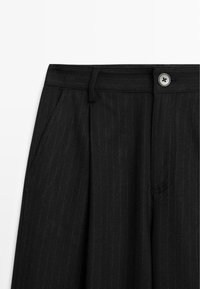 Pantalons noirs à rayures épaisses avec fermeture à bouton, design plissé, deux poches avant et coupe ajustée. Le tissu semble texturé.