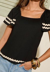 Černý krátký rukáv top s balónovými rukávy a čtvercovým výstřihem, s bílým vlnitým lemem na spodním okraji a okrajích rukávů.