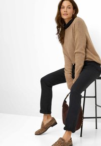 Beiger, flauschiger Pullover, schwarze Jeans, Loafers im Leopardenmuster und eine braune Handtasche. Modell sitzt auf einem schwarzen Metallhocker vor einem weißen Hintergrund.