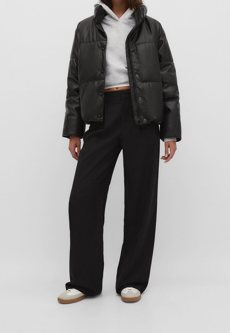 Personne portant une doudoune noire par-dessus un sweat à capuche court gris clair, un pantalon large noir et des baskets blanches avec des semelles marron.