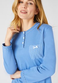 Maglietta a maniche lunghe di colore blu realizzata in cotone, con scollatura a bottoni, tasca a pois bianchi e cuciture decorative lungo il colletto e le maniche.