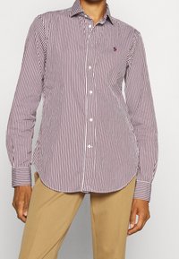 Camisa de botones de manga larga a rayas verticales en color burdeos y blanco, con un cuello puntiagudo y un pequeño logotipo en el pecho. Tejido suave.