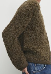 Bruine, gestructureerde trui met een grove breiwijze, voorzien van een ronde halslijn en geribbelde manchetten. Knopdetail zichtbaar aan de zijkant.