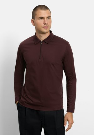 Mann mit kurzen Haaren, der ein burgunderfarbenes Poloshirt mit langen Ärmeln und Frontreißverschluss sowie schwarze Hose trägt, blickt zur Seite.