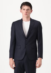 Jeune homme portant un costume bleu marine à rayures fines avec un t-shirt blanc à col rond, debout devant un fond blanc uni.