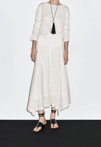 Valkoinen pitkähihaisen yläosa yhdistettynä liehuvaan, nilkkapituisen hameeseen. Mustat remmilliset sandaalit. Kaulakoru, jossa musta tupsuriipus.