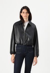 Giacca nera in pelle sintetica con chiusura a zip, ampio colletto e maniche a sbuffo, abbinata a jeans in denim scuro con tasche posteriori.