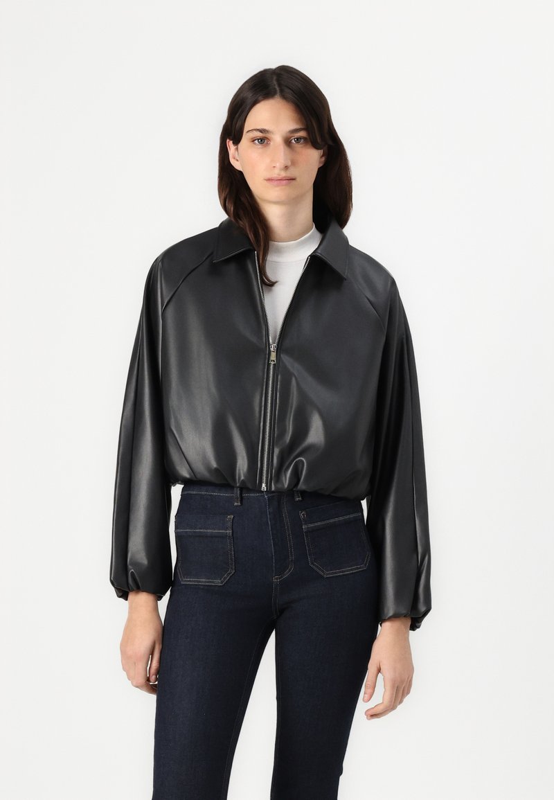 Giacca nera in pelle sintetica con chiusura a zip, ampio colletto e maniche a sbuffo, abbinata a jeans in denim scuro con tasche posteriori.