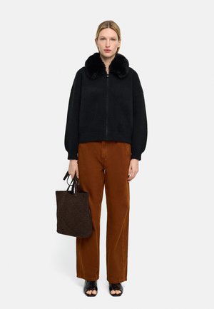 Kobieta ubrana w czarną kurtkę z futrzanym kołnierzem zapinaną na zamek, brązowe spodnie o szerokich nogawkach, czarne sandały z odkrytymi palcami, trzymająca ciemnobrązową teksturowaną torebkę, stojąca na białym tle.