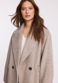 Beżowy, oversize'owy płaszcz w jodełkę z szerokim kołnierzem, dwoma przednimi kieszeniami i dwiema dużymi ciemnymi guzikami.