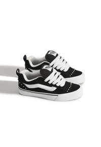 Zapatillas de ante en blanco y negro con un diseño grueso, cordones prominentes y tachuelas decorativas en el talón. Suelas de goma blancas.