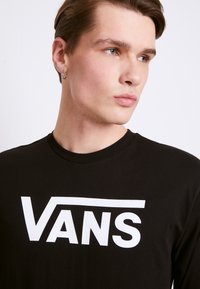 Sort langærmet T-shirt lavet af bomuld. Har et stort hvidt "VANS" logo foran. Rund halsudskæring, glat tekstur.