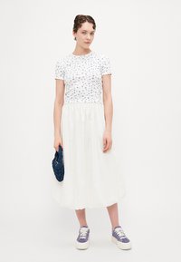 Witte bloemen top met korte mouwen, gecombineerd met een witte, vloeiende midi rok. Model draagt paarse sneakers en houdt een marineblauwe tas vast.