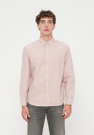 Joven con una camisa de manga larga a rayas blancas y rosa claro con botones y vaqueros grises, de pie frente a un fondo blanco liso.