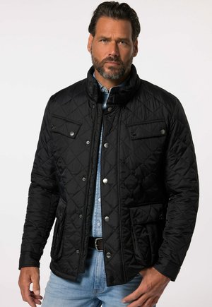 Sort quiltet jakke med høj krave, trykknaplukning og to forreste klaplommer. Bæres over en denimskjorte.