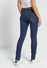 Jeans skinny de mezclilla azul oscuro con cintura alta, bolsillos traseros y un parche de cuero marrón en la cinturilla. Textura suave y diseño ajustado.