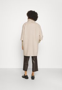 Suéter oversize beige con cuello alto, hombros caídos, puños acanalados y un sutil detalle de logo en la parte trasera. Combinado con pantalones de cuero marrón.
