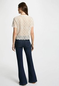 Top in pizzo con maniche corte in avorio, caratterizzato da un motivo geometrico e orlo ondulato, abbinato a jeans a zampa d'elefante blu scuro.