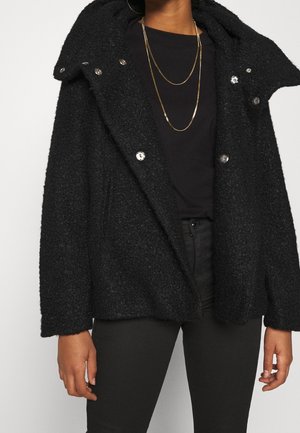 Persoon draagt een gestructureerd zwart jasje met een grote kraag, een zwart shirt, zwarte broek en gelaagde gouden kettingen.