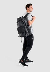 Mochila negra hecha de tela duradera con un bolsillo frontal de malla, múltiples compartimentos con cremallera y correas de hombro acolchadas.