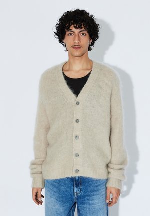 Béžový pletený cardigan z chlupatého materiálu s výstřihem do V, šesti šedými knoflíky a žebrovanými manžetami, v kombinaci s modrými džínami.