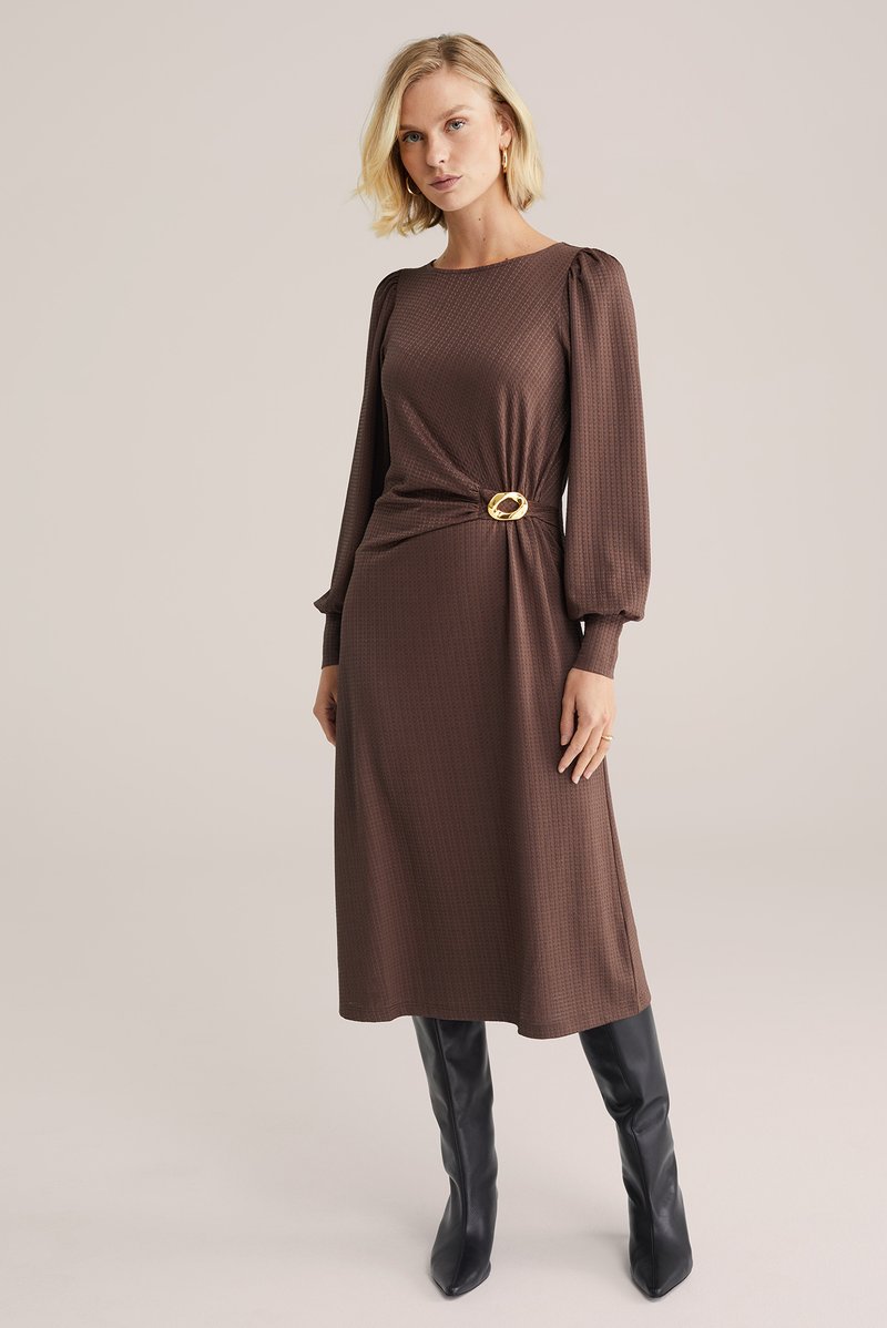 Brązowa, teksturowana sukienka midi z długimi bufami na rękawach i złotym okrągłym paskiem w talii, zestawiona z czarnymi kozakami za kolano.