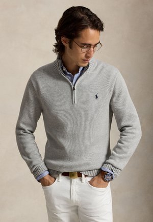 Un hombre con gafas luce un jersey gris con cremallera corta sobre una camisa de rayas y unos pantalones blancos, de pie y con las manos en los bolsillos.