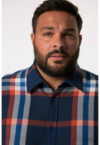 Camisa de cuadros en navy, rojo, azul y gris. Presenta un collar con puntas, tapeta de botones y una textura suave con un patrón de cuadros visible.