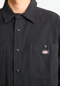 Black button-up shirt made of sturdy fabric. Features a collar, two chest pockets, and a logo patch on the left pocket. Buttons are black.