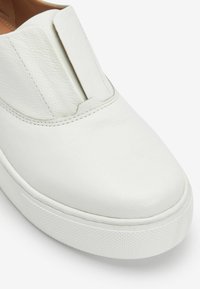 Chaussure slip-on blanche en cuir avec un dessus texturé et un design minimaliste, dotée d'un profil bas et d'une semelle en caoutchouc.