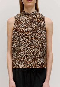 Ärmelloses Top mit Leopardenmuster und hohem Kragen, mit glatter Textur, beigem Grund und schwarzen sowie braunen Flecken. Kombiniert mit schwarzen Hosen.