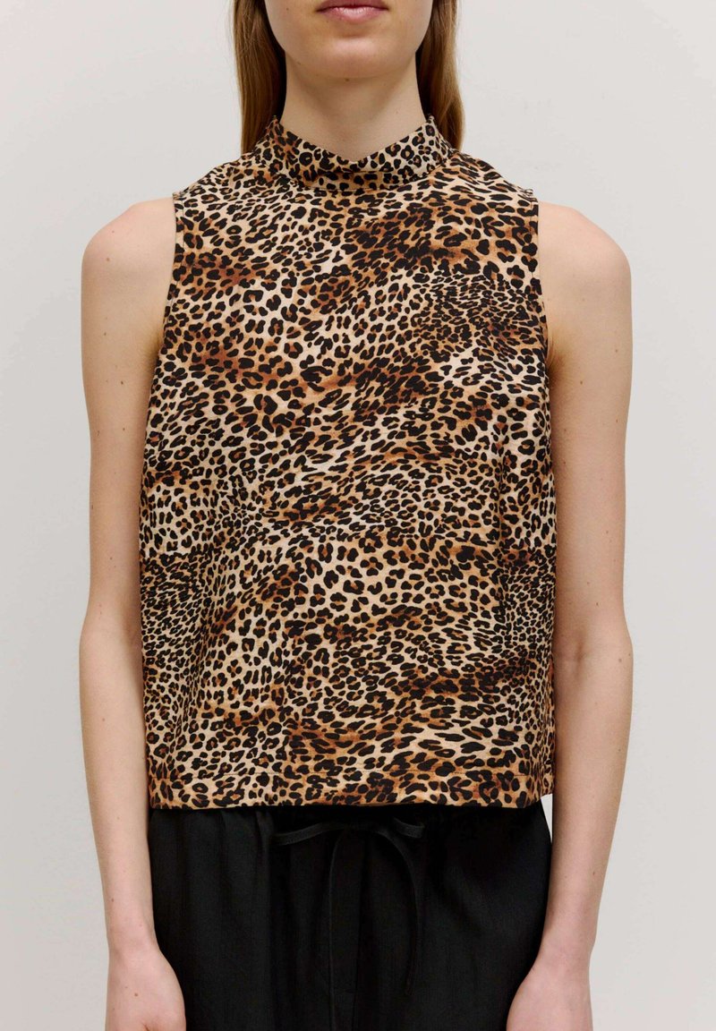 Ärmelloses Top mit Leopardenmuster und hohem Kragen, mit glatter Textur, beigem Grund und schwarzen sowie braunen Flecken. Kombiniert mit schwarzen Hosen.