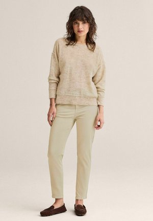 Kvinde med krøllet mørkt hår iført en beige sweater, lysebeige smalle bukser og mørkebrune mokkasin-sko, stående foran en ensfarvet baggrund.