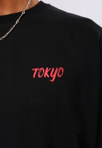 Μαύρο πουκάμισο με κόκκινο κείμενο "TOKYO" στο στήθος, φορεμένο με ασημένιο κολιέ αλυσίδα που είναι ορατό στον λαιμό.