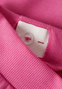 Tela rosa con una textura acanalada. La etiqueta blanca presenta un botón circular y una pequeña abertura con detalles de costura.