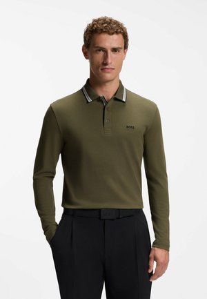 Uomo che indossa una polo a maniche lunghe verde oliva con colletto nero e bianco, pantaloni neri e cintura nera, in piedi su sfondo bianco.