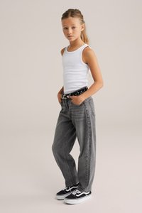 Camiseta sin mangas blanca, cinturón negro con tachuelas, jeans grises holgados y zapatillas negras con acentos blancos. La modelo está de pie con las manos en los bolsillos.