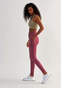 Leggings taille haute, ajustés, de couleur rose profond avec des poches latérales, associés à un soutien-gorge de sport olive clair et des chaussures de sport blanches.