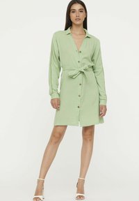 Robe boutonnée vert clair avec ceinture à la taille, manches longues et tissu texturé. Présente des boutons ronds et une longueur au genou.