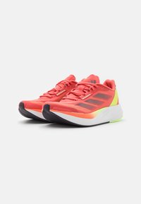 Svijetlucave koraljne trkaće tenisice s tri tamnosive pruge, teksturiranom mrežastom gornjištu, podstavljenim ovratnikom i bijelim potplatom s neonsko žutim naglascima.