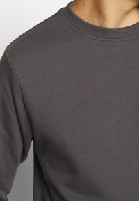 Gros plan d'une personne portant un sweat-shirt à col rond gris foncé uni, montrant l'épaule, le haut de la poitrine et la zone du cou.