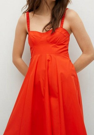 Femme portant une robe sans manches rouge vif, ajustée au niveau du buste avec de fines bretelles froncées et une jupe évasée, debout devant un fond uni.