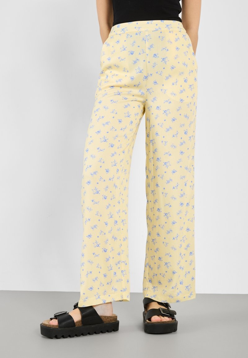Pantalon jaune à jambes larges avec un petit imprimé floral bleu porté avec des sandales noires et un haut noir.