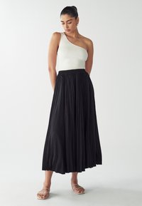 Black pleated maxi skirt, elastic waist, paired with a ribbed white one-shoulder top. Simple flat sandals complete the look.