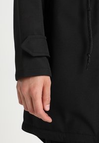 Abito in tessuto nero con una texture liscia, caratterizzato da un polsino decorato con un accento a forma di diamante e un cordino regolabile.