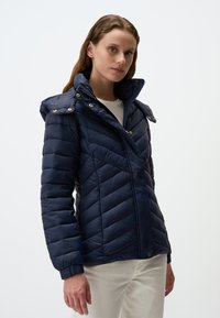 Giacca imbottita blu navy con trapunte a V, cappuccio rimovibile e dettagli in metallo dorato. Tasche con zip su entrambi i lati.