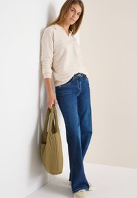 Beige Pullover mit Kragen, blaue Schlagjeans und eine grüne, gepolsterte Tragetasche. Slim Fit mit strukturiertem Strick, Modell lehnt an einer Wand.