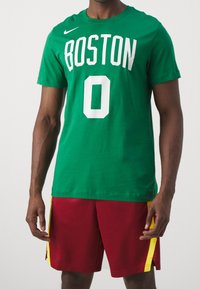 Man in een groene Boston t-shirt met het nummer 0 en rode sportshorts met gele strepen, staand tegen een effen achtergrond.